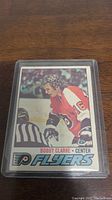 Front view of 1977-79 OPC Bobby Clarke hockey card featuring Bobby Clarke in a red Flyers jersey, number 16, facing a referee on the ice.