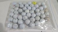 Full view of 50 used golf balls in a clear plastic bag on a white surface, mostly white balls with scuffs, including one yellow ball.