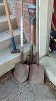 Three tools leaning against a brick wall and concrete steps: two rusted shovels and one sledgehammer with blue grip tape.