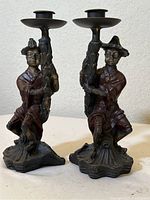 Pair of cast metal candlesticks with kneeling Asian figures holding the vertical stems topped with candle plates.