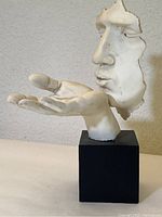 Front left angle of clay sculpture showing face blowing a kiss and open hand on black base.