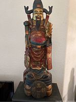 Front view of painted wood carving showing full statue, red and gold robes, base with large coin