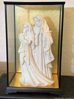 Front view of the white religious nativity figures of Mary, Joseph, and baby Jesus inside a sealed glass display case with black frame and gold backdrop.