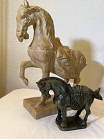 Two horse figurines shown together for scale and comparison. The larger beige composition horse with ornate details is positioned behind the smaller green stone carved horse figurine.