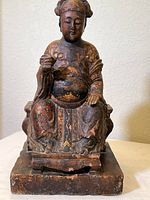 Full front view of the wooden carved figure seated with detailed hand-painted attire and a carved base.