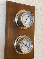 Wooden plaque with mounted thermometer and barometer dials, brass trim, measuring 7x16 inches.