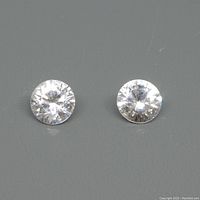 Two round, colourless natural zircon gemstones displayed on a dark surface showing their size, cut, and clarity.