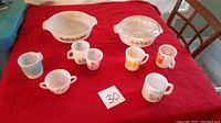 Photo of two white glass mixing bowls with colorful patterns and an assortment of seven small vintage mugs with various retro designs, all laid on a red cloth.