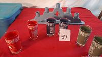 Four cylindrical collectible glasses in red, purple, and green with white Centennial designs displayed on a red cloth.