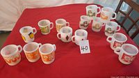 Photo showing 15 Fire King milk glass mugs with various colorful vintage designs, arranged on a red tablecloth.