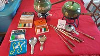 Overview of two green enamel fondue pots with wooden handles, stands, burners, and various utensils including fondue forks and small spoons in colorful boxes.