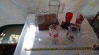 Wide view showing six glass ware items including pitchers with red floral designs, ribbed and rectangular vases, a glass item with a red stopper and print, and a clear cylindrical glass
