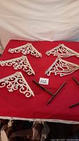 Eight white metal shelf brackets on red cloth, four scroll style and four floral style with some wear.