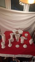Full lot of 13 white milk glass items arranged on a red cloth surface with white backdrop, includes various vases, bowls, and lidded containers.