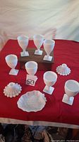 Full view of six milk glass wine glasses with square bases, two hobnail textured candle holders, and leaf-shaped fruit tray on a red cloth.