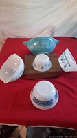 Five vintage mixing bowls displayed on a red fabric background: One large turquoise Pyrex bowl with white 'Friendship' pattern, four white Fire King bowls with blue floral or leaf designs.