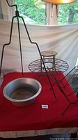 Full view of birdcage stand with hook and blue glass orb, round plant stand surface, metal basin below, and part of magazine rack on red cloth.