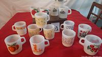 Overview of 15 white opaque Termocrisa mugs with various colorful vintage designs shown on red cloth table.