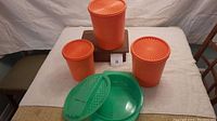 Photo of three orange Tupperware containers with lids and green salad bowl with accessories on a table.