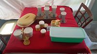 Photo shows entire collection of Tupperware items arranged on a red cloth, including spice containers, ice-cream holders, measuring spoons, and veggie holders.