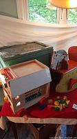 Wide view of the vintage wooden rocking horse, wooden activity house on wheels, and scattered colored wooden blocks and hammer on red tablecloth