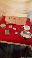 Shows all items in the lot arranged on a red tablecloth including the white tin container with 4 drawers, ceramic bird container, metal potato masher, porcelain bowl, metal baking pan, glass lidded container, pocket watch, and yellow-blue manual day timer calendar.