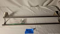 Metal towel rack with dual bars and mounting hardware in plastic bag, placed on white background.