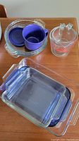 Assorted glassware items including two clear glass pie plates, a dark blue glass measuring cup, a clear glass measuring cup with red measurement markings, and two rectangular glass baking dishes with blue handles on a wood surface.