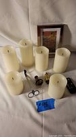 Six ivory battery operated candles arranged with a framed postcard, vintage brass whoopee whistle, candle wick trimmer, kaleidoscope, and remote control on white fabric background.