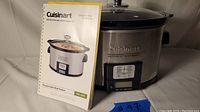 Photo showing Cuisinart programmable slow cooker with stainless steel body and black trim, digital control panel, and glass lid. Instruction and recipe booklet placed next to the slow cooker.