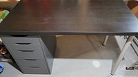 Full view of the IKEA desk showing dark wood surface, cabinet with drawers on left, and metal leg on right.