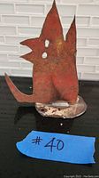 Photo shows a small metal statuette shaped like an abstract howling dog, painted rusty red with visible rust and chipped areas on a metal base.