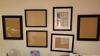 Six black picture frames arranged on a wall, some containing cardboard backing or printed paper, in excellent condition.