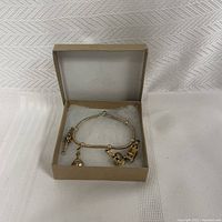 Gold-coloured charm bracelet with five hanging charms displayed inside a beige box on a white textured background.
