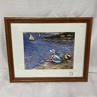 Framed original artwork featuring a girl sitting on the beach next to a colorful ball, sailboats and distant houses visible in the scene.
