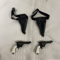 Two porcelain revolvers with white grips placed next to matching black holsters with straps laid flat on white fabric background.
