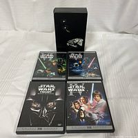 Four DVD cases displayed with covers facing up, including three Star Wars trilogy episodes and one bonus material disc, arranged in front of a black box with Star Wars space ships printed on it.
