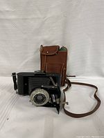 Vintage Kodak Brownie Flash Six-20 film camera with folding bellows extended, positioned next to brown leather carry case with strap and front pocket.
