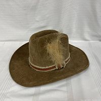 Front angled side view of a brown corduroy western style hat on a white background showing the wide flat brim and feather accent on striped band.