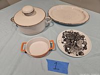 Four items shown: Stonehenge Midwinter casserole with lid, oval speckled plate, CB2 small plate with black decoration, small enameled crock with orange handles.