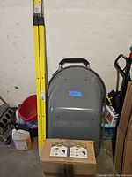 Side view showing the grey steel tray of the wheelbarrow leaning against a wall next to yellow steel handles and a boxed parts carton.