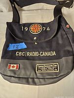 Replica messenger bag showing front view with 1974 embroidered text and CBC Radio Canada branding, canvas material upper and rubber bottom
