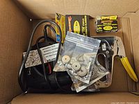 Photo showing manual staple tool with yellow handle, electric staple tool, boxes of staples and nails, and several small sealed bags with magnets and metal parts inside a cardboard box.