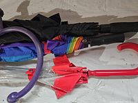 Top view of the purple curved Trigger Point massager alongside three large umbrellas: black, rainbow-colored, and clear umbrella with red handle.