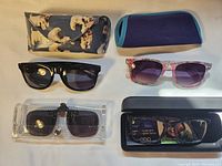 Two sunglasses, one clip-on sunglasses, one pair of prescription glasses in their cases displayed on a surface.