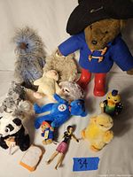 Photo showing Paddington Bear in blue coat, black hat with pin, red boots, and various stuffed animals including blue bird, yellow duck, panda, Gina Carano figure, and others.