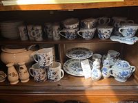 Photo showing upper and lower shelves displaying various blue and white china cups, saucers, plates, and a white soup tureen. Blue Danube designs visible with floral patterns.