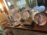 Wide angle shot showing the full grouping of collector plates, candlesticks, an Asian styled plate on stand, metal elephant figurines, and tiered decorative metal item