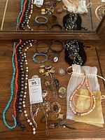 Top view of multiple costume jewelry items including beaded necklaces in coral, white, purple, pins, rings, and bracelets laid out on a wooden surface with two small drawstring pouches.