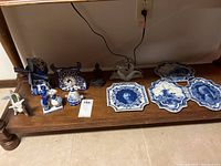 Full overview of the collection showing Delft plates, figurines, mosaic clock, Buddha statues, and a ceramic cow item on a wooden table.